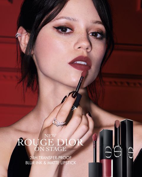 Una donna applica il rossetto "Rouge Dior" tenendo l'applicatore vicino alle labbra. Sono raffigurati tre tubetti di rossetto. Il testo pubblicizza un rossetto a lunga tenuta di 24 ore, che non sbava, sfuma delicatamente le labbra e offre un finish opaco.