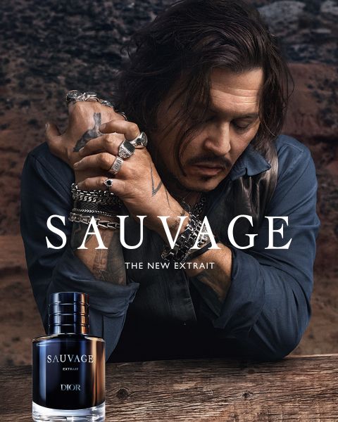 Un uomo con tatuaggi e gioielli si appoggia a un tavolo accanto a una bottiglia di profumo "Dior Sauvage Extrait" davanti a uno sfondo esterno accidentato.