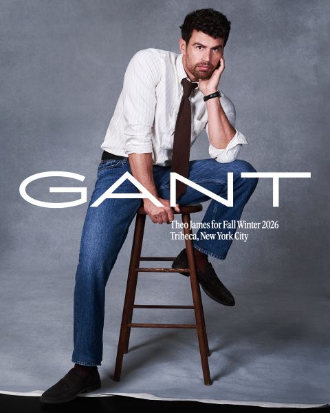 Un uomo in camicia a righe, cravatta e jeans posa su uno sgabello di legno davanti a uno sfondo grigio. Testo: "GANT, Theo James per Autunno/Inverno 2026, Tribeca, New York City.".