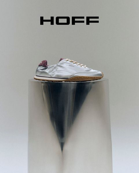 Una sneaker argentata con tacco viola si trova su un piedistallo riflettente sotto il logo "HOFF" su uno sfondo grigio chiaro.