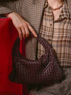 Una persona in un abito a quadri che tiene una borsa a mano intrecciata marrone scuro; sullo sfondo si vede parzialmente uno sfondo rosso.