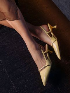 Una donna che indossa décolleté gialle a punta con cinturino è seduta su un pavimento di legno scuro; la sua gonna marrone trasparente è parzialmente visibile.