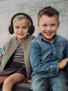 Due bambini piccoli sono seduti uno accanto all'altro, una bambina con paraorecchie e una giacca e un bambino con una camicia e jeans di jeans; entrambi sorridono alla telecamera.
