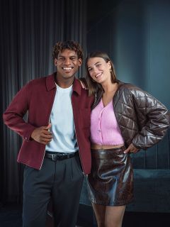 Un uomo sorridente con una giacca bordeaux sta accanto a una donna con un top rosa e una gonna marrone; entrambi posano in un interno davanti a uno sfondo scuro.