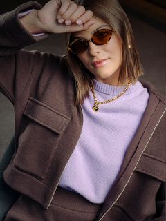 Una donna con occhiali da sole marroni, giacca e maglione color lavanda appoggia il braccio sulla fronte; indossa gioielli d'oro e ha un'espressione sicura di sé.