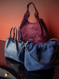 Tre borse a mano eleganti - una in camoscio bordeaux, una in pelle blu navy e una in camoscio blu navy con tracolla a catena - sono esposte su uno sfondo marrone rossiccio.