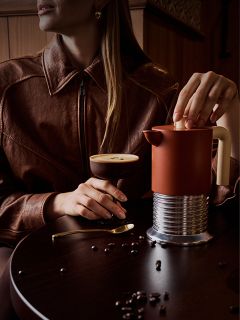 Una donna con una giacca marrone siede a un tavolo, sul quale ci sono un cocktail al caffè, una French Press e chicchi di caffè sparsi.