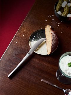 Un coltello su un piatto con una fetta di pane, un po' di sale e cetriolini sottaceto, e una bevanda cremosa accanto su un tavolo di legno.