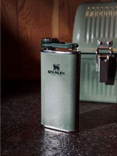 Su un piano di lavoro scuro c'è una borraccia termica Stanley verde, sullo sfondo si vede un portapranzo verde abbinato davanti a una parete di legno.