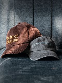 Due berretti su una poltrona di stoffa blu; uno marrone con la scritta „The Art of Being Late“, l'altro in nero sbiadito con una scritta più piccola e difficilmente leggibile.