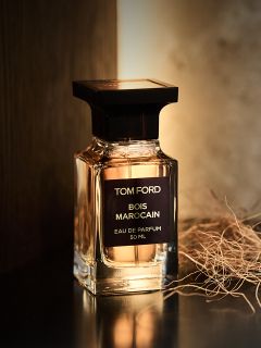 Un flacone di profumo „Bois Marocain“ di Tom Ford si trova su una superficie scura accanto a erba secca, mentre una luce calda lo illumina da dietro.