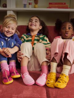Tre piccoli bambini siedono su un divano rosso, sorridono e ridono; ciascuno indossa abiti colorati e scarpe diverse e vivaci.