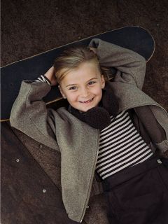 Un bambino sorridente con una maglietta a righe e una giacca è sdraiato su uno skateboard, le mani incrociate dietro la testa, e guarda in camera.