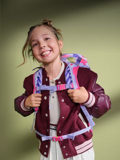 Una ragazza sorridente con uno zaino e una giacca bordeaux si trova davanti a uno sfondo verde, apparendo allegra e pronta per la scuola.
