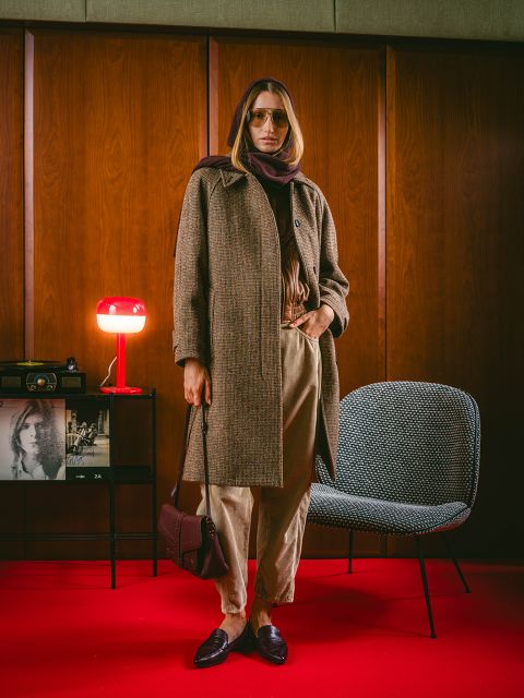 Una persona con un cappotto marrone e una sciarpa è in piedi in una stanza su un tappeto rosso, sullo sfondo si vedono decorazioni vintage, una sedia e una lampada rossa.
