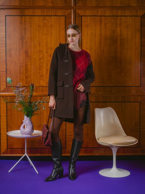 Una donna con un cappotto scuro e stivali è in piedi accanto a una sedia bianca e un tavolo con fiori davanti a una parete di legno e un pavimento viola.