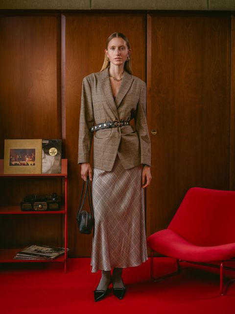 Una donna in blazer e gonna a quadri si trova in una stanza retrò con pareti rivestite in legno, un tappeto rosso, una sedia rossa e oggetti su uno scaffale rosso.