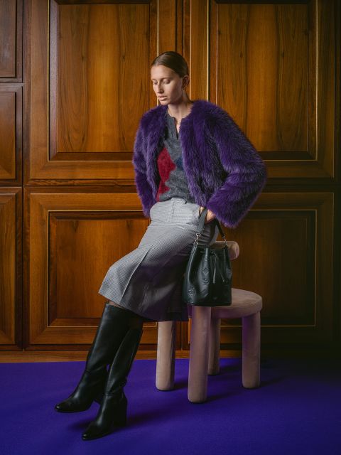 Una donna con una giacca di pelliccia viola, una gonna grigia e stivali neri è seduta su uno sgabello con una borsa nera davanti a pannelli di legno su un tappeto viola.
