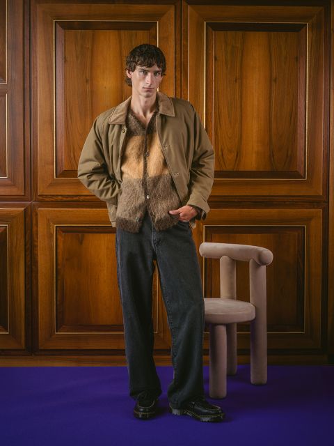 Un uomo con una giacca marrone, una camicia pelosa e jeans neri è in una stanza accanto a una poltrona beige, le cui pareti sono rivestite di pannelli di legno e su cui è posato un tappeto viola.