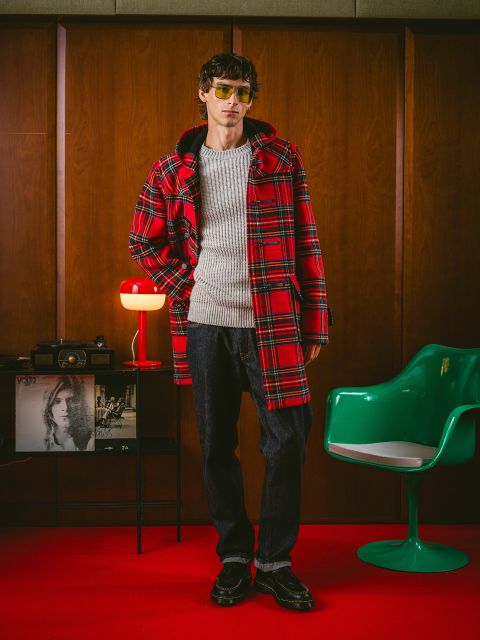 Una persona con un cappotto a quadri rosso, un maglione grigio, jeans scuri e occhiali da sole si trova in una stanza in stile retrò con pannelli in legno, una sedia verde e una lampada rossa.