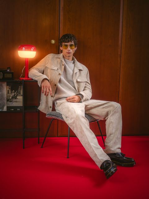 Un uomo vestito di beige è seduto su una sedia su un tappeto rosso, dietro di lui ci sono una lampada rossa e una parete di legno. Indossa occhiali da sole e scarpe nere.
