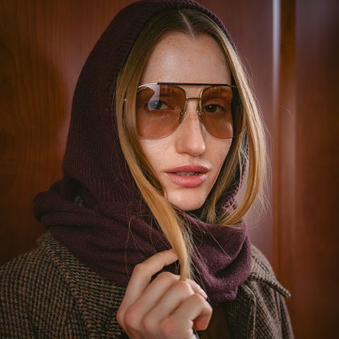 Una donna con grandi occhiali da sole, un cappuccio sciarpa bordeaux e un cappotto a quadri marrone si trova di fronte a uno sfondo di legno.