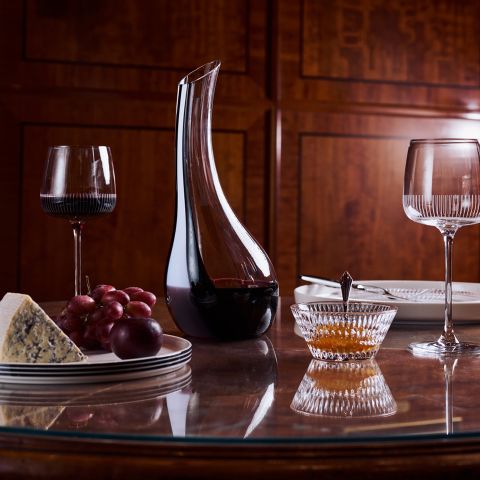 Una caraffa di vino, due bicchieri, formaggio, uva e una ciotola di miele su un tavolo di legno lucido, con dei piatti visibili sullo sfondo.
