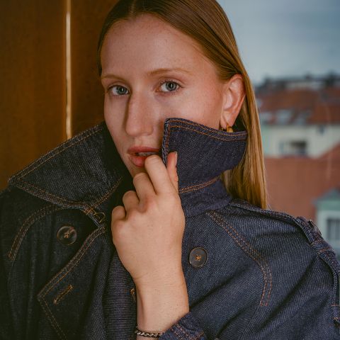 Una donna dalla pelle chiara e i capelli lisci, che indossa una giacca di jeans scura con il colletto alzato, in piedi davanti a una finestra, con edifici visibili sullo sfondo.