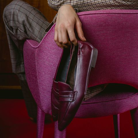 Una persona in pantaloni a quadri è seduta su una sedia rosa e tiene un mocassino bordeaux sullo schienale della sedia.