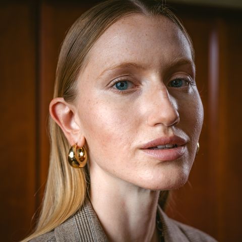 Una donna con capelli biondi lisci, orecchini a cerchio dorati e trucco discreto guarda la telecamera mentre si trova di fronte a uno sfondo di legno.