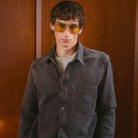 Un giovane uomo con capelli castani ondulati, che indossa occhiali da sole gialli, una camicia di jeans grigia e un maglione bianco, in piedi davanti a uno sfondo con pannelli di legno.