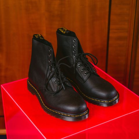 Un paio di stivali in pelle nera con lacci e cuciture decorative gialle sono posizionati su un lucido espositore rosso davanti a uno sfondo di legno.