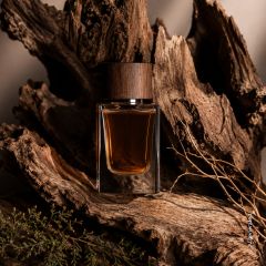 Un flacone di profumo in vetro con un tappo di legno si trova in mezzo a corteccia d'albero ruvida e rami secchi, creando un'atmosfera rustica e naturale.