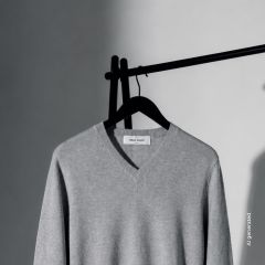 Un maglione grigio chiaro con scollo a V è appeso a una gruccia nera davanti a uno sfondo semplice, in parte in ombra, in parte alla luce.