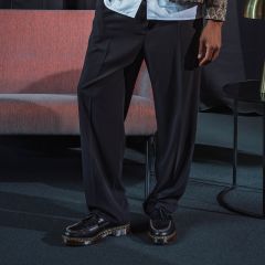 Una persona si trova in un ambiente interno e indossa pantaloni neri a gamba larga e scarpe nere con plateau; accanto a lei ci sono un divano rosa e un tavolino rotondo.