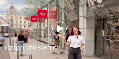 Eine Frau winkt vor einem K&Ö-Geschäft auf einer belebten Stadtstraße mit Passanten und roten Schildern. Der Text lautet: „Scopri chi siamo.“.