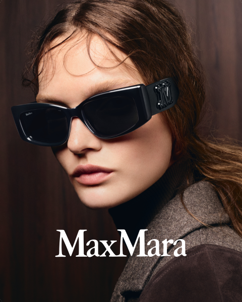 Una donna con grandi occhiali da sole neri e un cappotto marrone su uno sfondo di legno scuro, con il logo "MaxMara" visibile in basso.