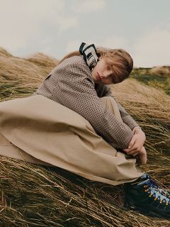 Una persona con un cappotto a quadri e una gonna lunga è seduta all'aperto nell'erba alta, guardando pensierosa la fotocamera.