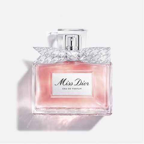 Una bottiglia di vetro trasparente di "Miss Dior Eau de Parfum", riempita con liquido rosa e un fiocco argentato e decorato intorno al collo della bottiglia, su uno sfondo bianco semplice.