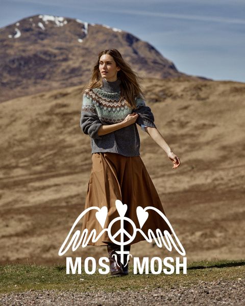 Una donna con un maglione a fantasia e una gonna marrone è in piedi all'aperto, con colline e una montagna visibili dietro di lei. Il logo "MOSS MOSH" è visibile nella parte inferiore dell'immagine.