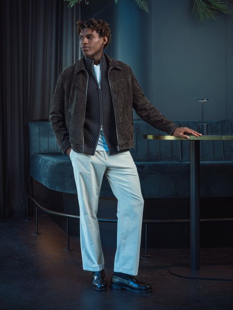 Un uomo con una giacca marrone e pantaloni bianchi sta accanto a un tavolo rotondo in una lounge moderna e poco illuminata, con sedute e tende blu scuro.