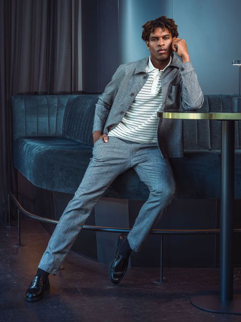 Un uomo in abito grigio e camicia a righe si appoggia a un tavolo rotondo in una lounge moderna con poltrone in velluto blu scuro.