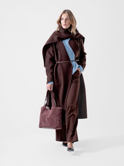 Una donna con un cappotto marrone scuro e pantaloni marrone scuro, oltre a un maglione blu, che tiene in mano una borsa marrone abbinata e cammina con sicurezza davanti a uno sfondo bianco semplice.