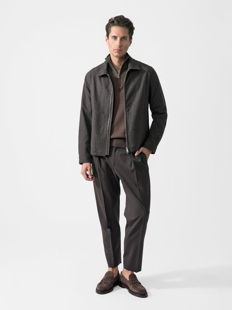 Un uomo che indossa una giacca a righe scure, un maglione marrone, pantaloni scuri e mocassini marroni, sta in piedi davanti a uno sfondo bianco semplice.