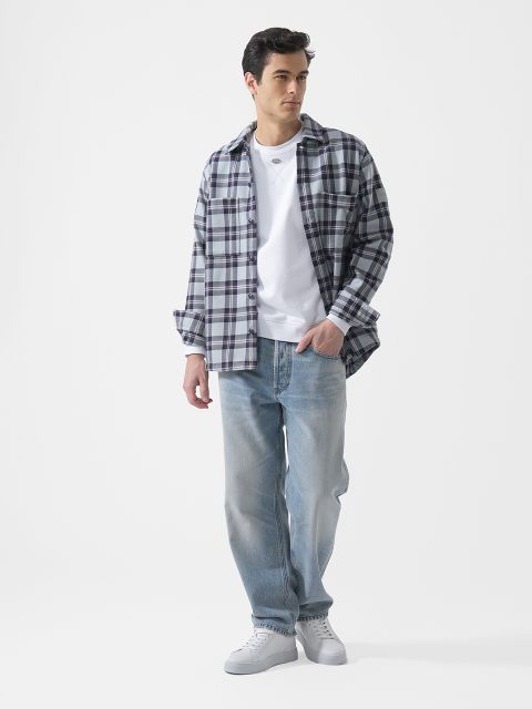 Un uomo che indossa una maglietta bianca, una camicia a quadri, jeans azzurri e scarpe da ginnastica bianche sta in piedi davanti a uno sfondo bianco semplice.