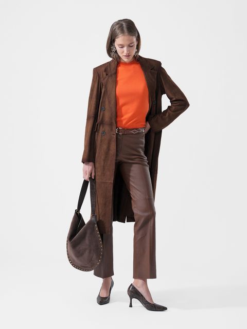 Una donna con un cappotto marrone, un top arancione, pantaloni marroni e décolleté tiene una borsa marrone e si trova davanti a uno sfondo bianco semplice.