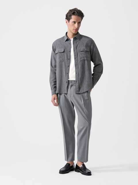 Un uomo che indossa una camicia grigia abbottonata, una maglietta bianca, pantaloni eleganti grigi e mocassini neri, in piedi davanti a uno sfondo bianco semplice.