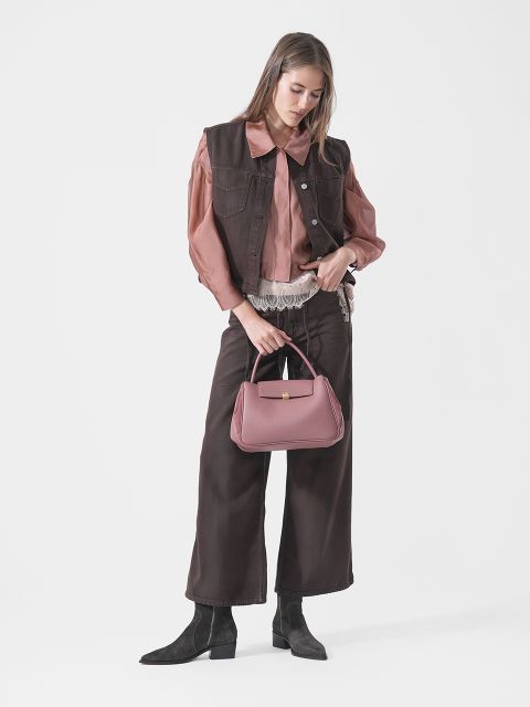 Una donna con gilet e pantaloni marroni, camicetta rosa e stivali neri tiene una borsa rosa e si trova davanti a uno sfondo bianco semplice.