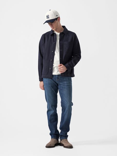 Un uomo che indossa una giacca blu navy, una maglietta bianca, jeans blu, stivali grigi e un cappellino da baseball bianco con la scritta NY, si trova davanti a uno sfondo bianco semplice.
