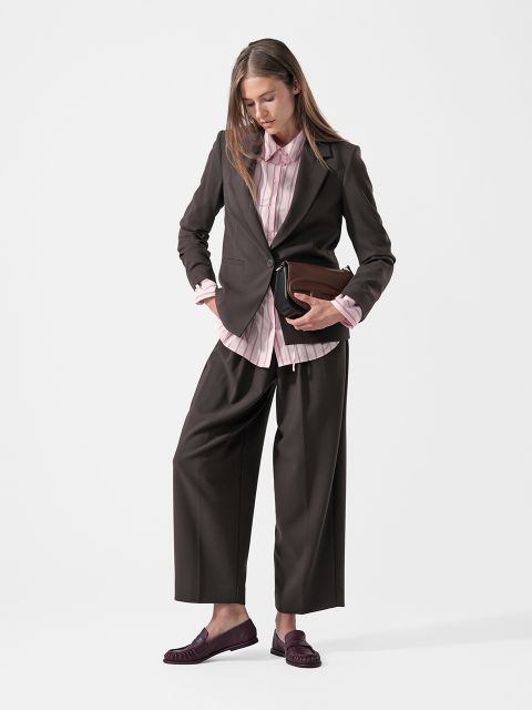 Una donna in tailleur pantalone scuro, camicia rosa e mocassini tiene in mano una pochette marrone, in piedi davanti a uno sfondo bianco semplice e guarda in basso.